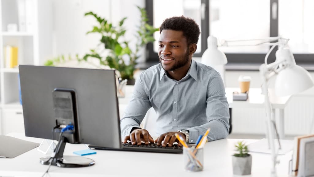 A man in a light blue shirt sits at a desk typing on a keyboard and looking at a computer monitor with the SimplePractice platform open in a bright, modern office workspace.